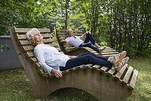 In einem Park liegen auf hözernen, geschwungenen Liegen eine Frau und ein Mann, beide sind etwas älter und haben weiße Haare.