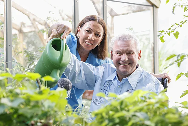 Eine Junge Frau hilft einem älteren Herrn eine Gießkanne zu halten und Blumen zu gießen. Beide sehen sehr zufrieden aus.