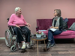 Eine Frau sitzt auf einem lila Sofa und unterhält sich mit einer älteren Dame, die in einem Rollstuhl sitzt. Der Raum sieht sehr gemütlich aus.