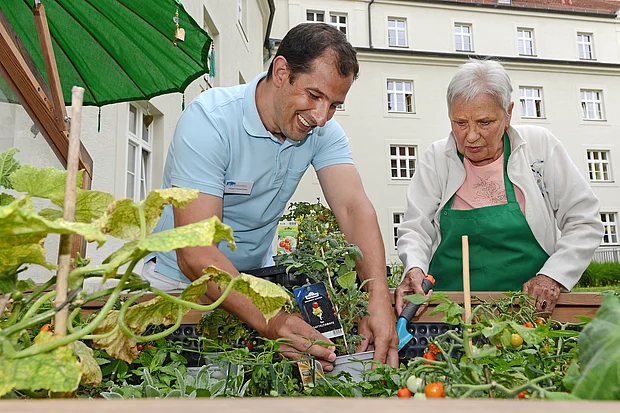 Ein Pfleger steht mit einem weiblichen Gast aus der Tagespflege an einem Hochbeet.