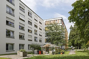 DAS Bild zeigt zwei moderne Gebäude mit einem kleinen Gartenhaus davor und hohen grünen Bäumen am Rand
