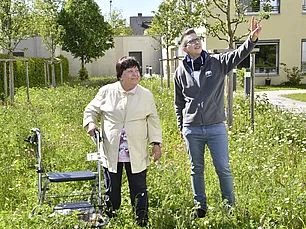 Eine junge Frau hebt die hand und zeigt in den Himmel. Neben ihr stehgt eine ältere DAme mit Rollator, sie stehen auf einer grünen Wiese.