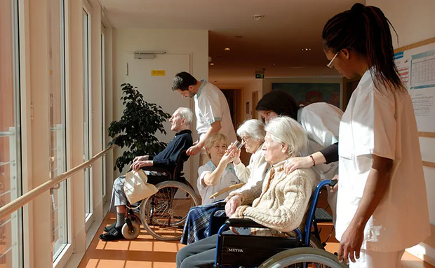 in einer Reihe sitzen viele ältere Menschen in der Sonne in Rollstühlen, um sie herum sind Pflegekräfte.