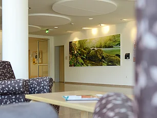 Ein heller freundlicher Flur mit schönen Bildern und großen flachen runden Lampen an der Decke ist zu sehen, außerdem gemütliche Sessel und Sofa und ein Sofatisch.