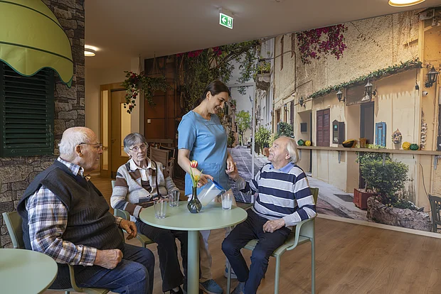 Eine junge Pflegerin steht neben einem älteren Herrn, der an einem Tisch sitzt. Im Hintergrund sieht man ein italienisches Steinhaus, zwei weitere ältere Personen sitzen am Tisch, im Hintergrund sieht man Pflanzen.
