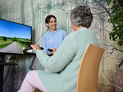 Eine ältere Dame sitzt auf einem Ergometer, eine Pflegerin steht daneben.