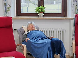 Eine ältere Dame liegt in einem Sessel, bei dem man die Liegefläche flach stellen kann. sie ist mit einer Decke zugedeckt und ruht.
