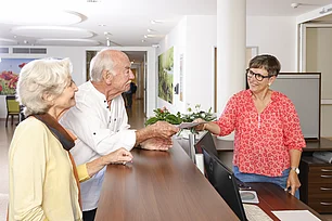 An einer Rezepotion steht eine Frau mit kurzen dunklen Haaren und reicht einem älteren Herrn etwas. Neben diesem steht eine ältere Frau. 