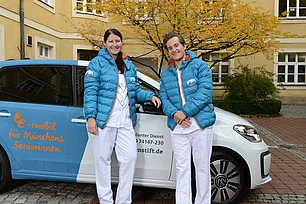 Eine junge Frau und ein junger Mann in Dienstkleidung lehnen an einem Auto des Ambulanten PFlegediensts.