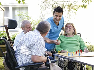 Drei Menschen in Rollstühlen sitzen im Garten, ein Brettspiel vor ihnen, sie lachen fröhlich. Ein Pfleger beugt sich zu ihnen und lächelt.
