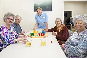Gäste der Tagespflege machen ein Gesellschaftsspiel, eine Pflegerin steht dabei. Alle haben Spaß.
