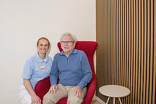 Eine Pflegerin sitzt in der Hocke neben einem älteren Herrn, der in einem Sessel sitzt und in die Kamera lacht.
