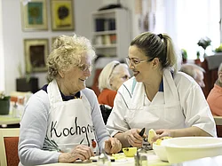 Eine Pflegerin der Tagespflege kocht mit einer älteren Dame, sie haben beide Schrüzen an und lächeln.