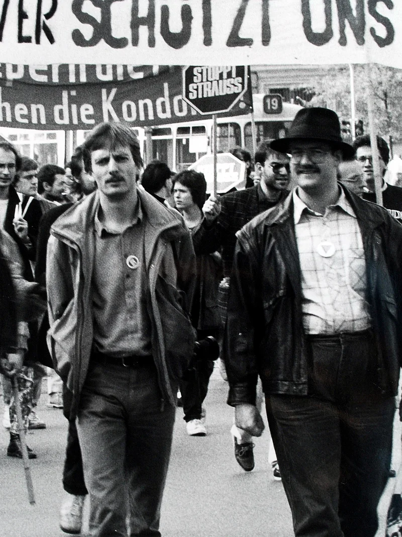 Eine schwarz-Weiß Fotografie von einer Demonstration, zwei Männer, dahinter Plakate, die man nur teilweise lesen kann.
