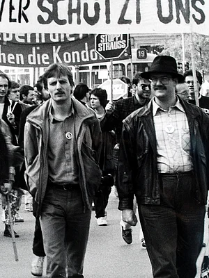 Eine schwarz-Weiß Fotografie von einer Demonstration, zwei Männer, dahinter Plakate, die man nur teilweise lesen kann.