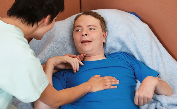 Ein junger Mann liegt lächelnd Im Bett, eine Pflegerin beugt sich über ihn.